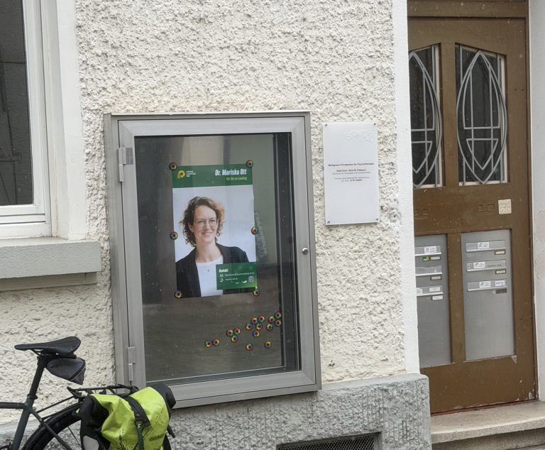Schaukasten mit einem Plakat der Grünen Landtagsabgeordneten und rechts daneben die Eingangstür; links vom Schaukasten sieht man ein Fahrrad mit Satteltasche