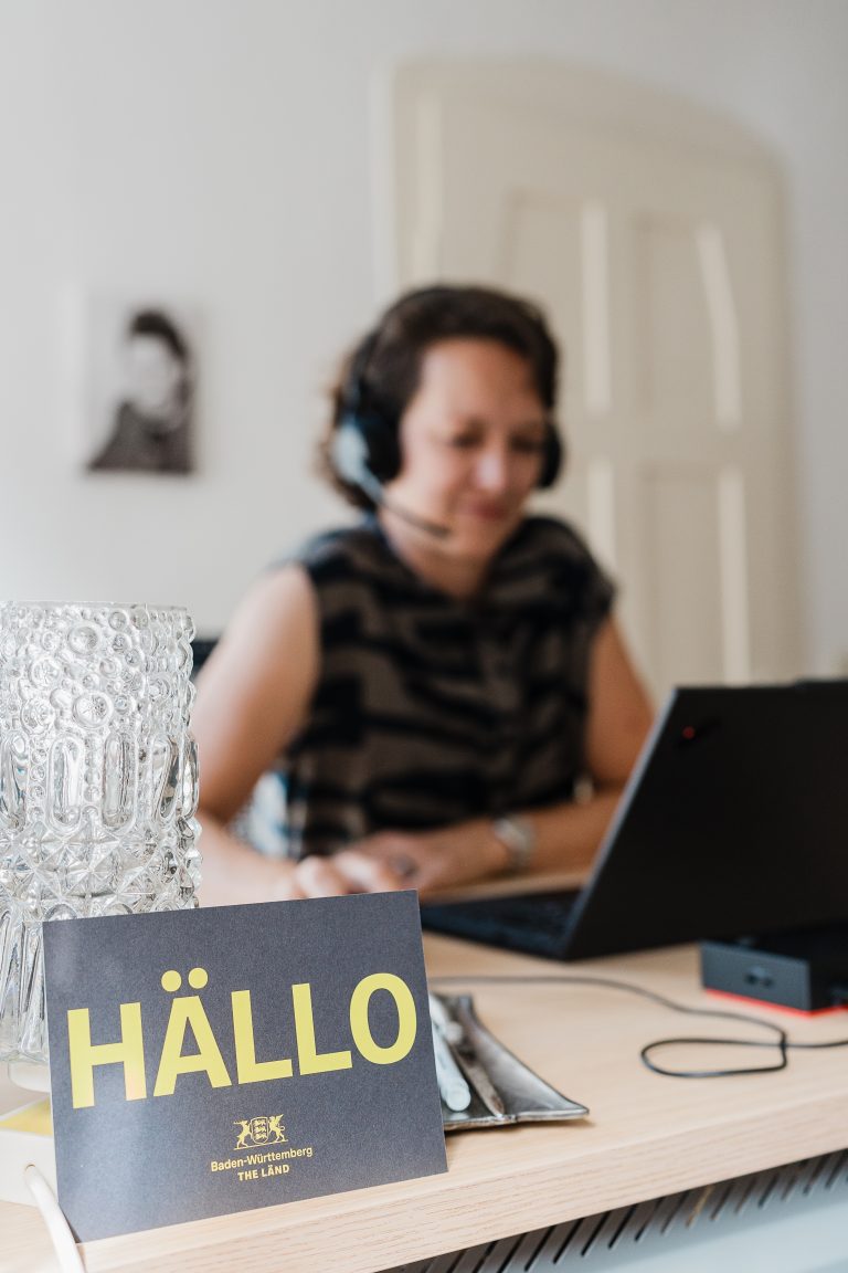 Mariska sitzt am Schreibtisch. Vorne ist eine Postkarte mit Aufschrift HÄLLO zu sehen.