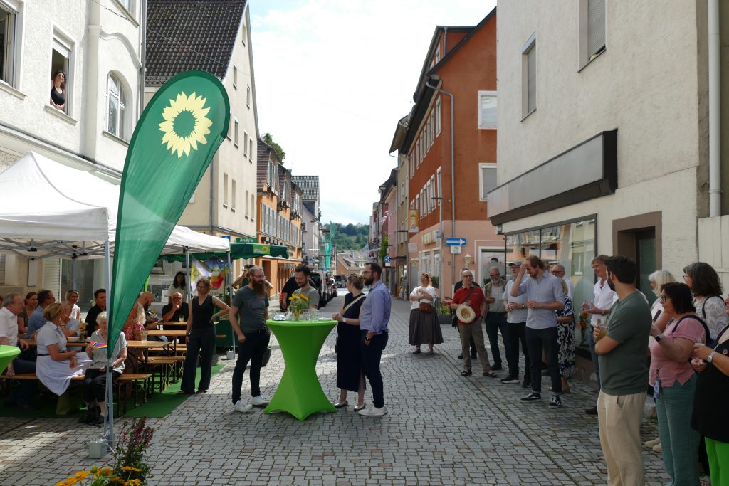 Blick auf die Straße vor dem Gebäude mit grüner Fahne, grünem Stehtisch und vielen Gästen